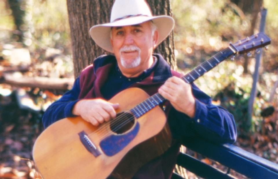 Man in hat leaning against a tree and playing the guitar